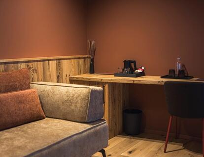 Gemütliche Sitzecke im Hotelzimmer mit gepolstertem Sofa, Schreibtisch aus Holz, Stuhl und warmer indirekter Beleuchtung.