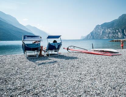 Two sun loungers and a windsurf board with sail on a pebble beach at Lake Garda, with views of the water and surrounding mountain landscape.