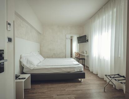 Modernes Zimmer im Hotel Villa Alberta mit Doppelbett, weißer Bettwäsche, Holzboden, Schreibtisch mit Stühlen, Fernseher und großen Fenstern mit Vorhängen.
