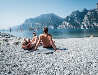 Due persone si rilassano su una spiaggia di ghiaia sul Lago di Garda, con vista sull’acqua turchese e sul paesaggio montano circostante.