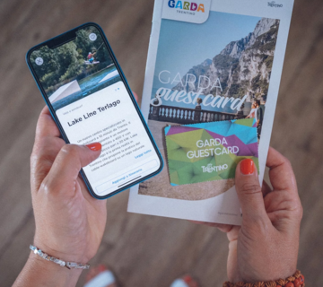 Person hält ein Smartphone und eine touristische Informationsbroschüre der Garda Guestcard in den Händen.