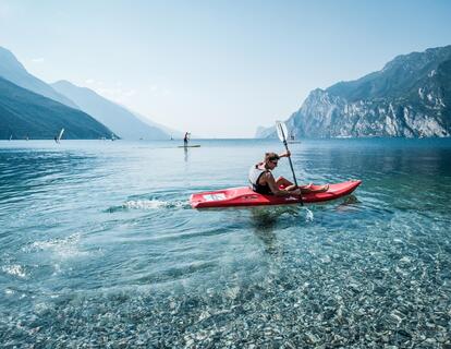 Persona a bordo di un kayak rosso sulle acque turchesi del Lago di Garda, con vista sul paesaggio montano circostante.