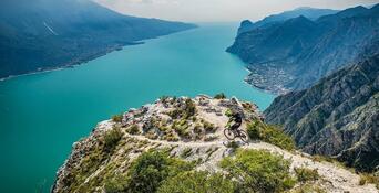 Mountain biker su un punto panoramico roccioso con vista sul Lago di Garda dalle acque turchesi e sul paesaggio montano circostante.