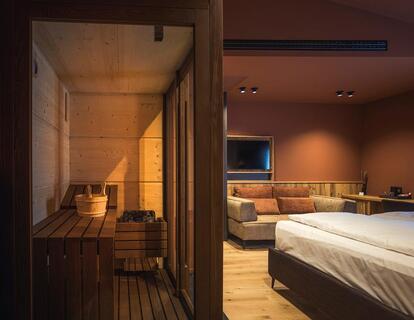 Modernes Hotelzimmer mit Doppelbett, Holzwänden, warmen Lichtakzenten, Fernseher und Blick durch eine Glastür in die angrenzende Sauna.