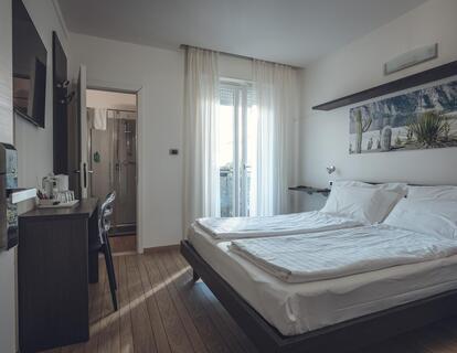 Zimmer im Hotel Villa Alberta mit großem Doppelbett, floralem Wandbild, Schreibtisch, Holzboden, Blick ins Badezimmer und auf den Balkon.