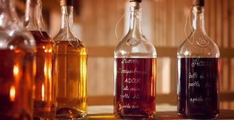 Diverse bottiglie di vetro contenenti distillati dalle tonalità dorate e ambrate su un tavolo di legno.