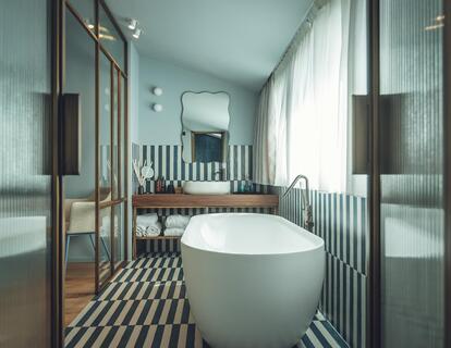 Bagno della Suite Aura con vasca da bagno bianca freestanding, lavabo, grande specchio, pavimento piastrellato in bianco e nero e finestre.