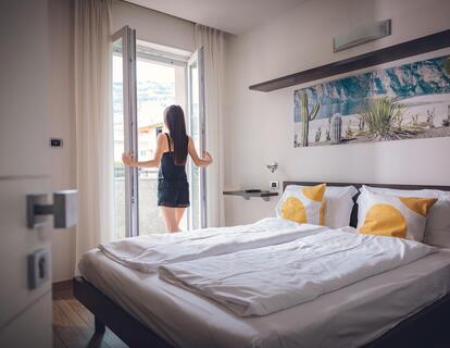 Frau steht vor der Balkontür im Hotelzimmer mit Doppelbett, gemusterten Kissen und großem Wandbild über dem Bett.