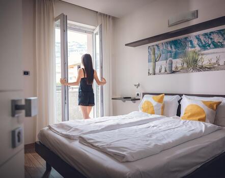 Frau steht vor der Balkontür im Hotelzimmer mit Doppelbett, gemusterten Kissen und großem Wandbild über dem Bett.
