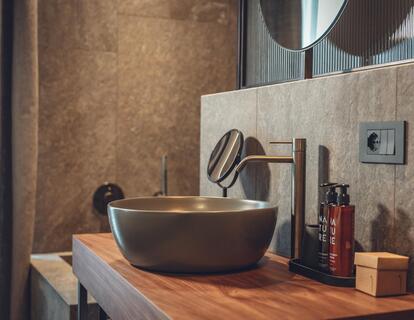 Close-up of a dark countertop washbasin on a wooden shelf with a round mirror and toiletries.