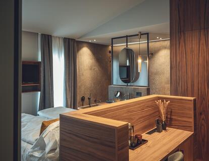 Modernes Hotelzimmer mit Doppelbett, Holzmöbeln, warmen Lichtakzenten und Blick ins Badezimmer mit großem Spiegel.