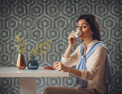 Donna seduta a un tavolo mentre beve da una tazza davanti a una parete con motivi geometrici neri e blu.