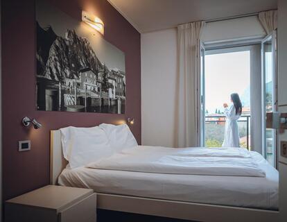 Helles Hotelzimmer mit Doppelbett, Nachttisch, großem Schwarz-Weiß-Bild über dem Bett und einer Frau auf dem Balkon.