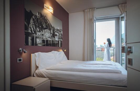 Helles Hotelzimmer mit Doppelbett, Nachttisch, großem Schwarz-Weiß-Bild über dem Bett und einer Frau auf dem Balkon.