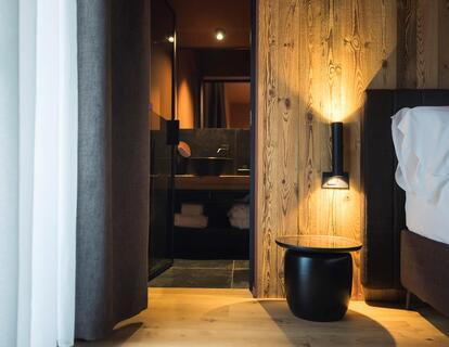 Modernes Hotelzimmer mit Doppelbett, Holzwänden, Nachttisch mit Lampe und Blick durch den Flur ins angrenzende Badezimmer.