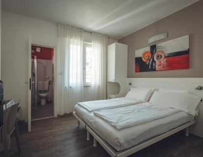 Modernes Zimmer im Hotel Villa Alberta mit Doppelbett, weißer Bettwäsche, großem buntem Wandbild, Holzboden, Fenster und Blick ins Badezimmer.