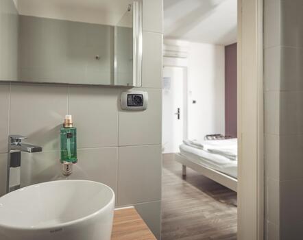 Modernes Badezimmer mit weißem Waschbecken, Spiegel und Pflegeprodukten sowie Blick ins Zimmer.