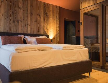 Camera d'hotel moderna con letto matrimoniale, parete in legno rustico, calde luci a parete e sauna con porte a specchio.