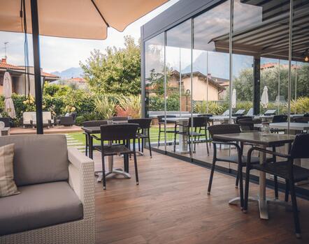 Hotelterrasse mit gepolsterten Sitzmöbeln, Tischen, Stühlen und Sonnenschirmen vor einer großen Glasfront.