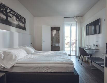 Modernes Zimmer im Hotel Villa Alberta mit Doppelbett, Holzboden, Wandbildern, Schreibtisch mit Stuhl, Fernseher und Balkontür.