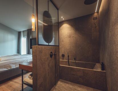 Bathroom in the Atlas Suite with dark stone tiles, walk-in shower, bathtub and a view of the double bed.