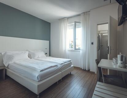 Modernes Zimmer im Hotel Villa Alberta mit Doppelbett, weißer Bettwäsche, weißem Kopfteil, Holzboden, Schreibtisch mit Stuhl, Fernseher und Fenster.