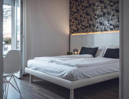 Modernes Hotelzimmer mit Doppelbett, weißer Bettwäsche, gemusterter Wandtapete und Balkon.