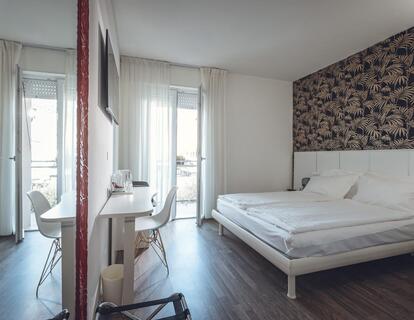 Modernes Zimmer im Hotel Villa Alberta mit Doppelbett, weißer Bettwäsche, weißem Kopfteil, floralem Wandbild, Holzboden, Schreibtisch mit Stuhl, Fernseher und Balkontür.