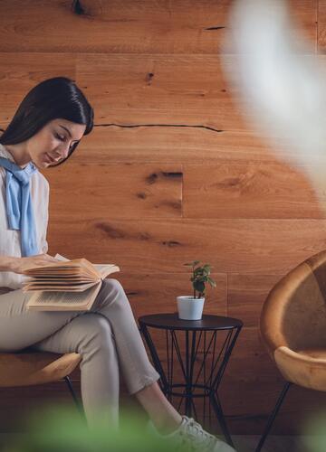 Donna seduta su una poltrona imbottita mentre legge un libro davanti a una parete in legno.