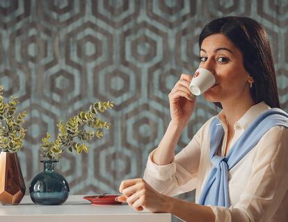 Donna seduta a un tavolo mentre beve da una tazza davanti a una parete con motivi geometrici neri e blu.