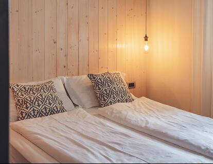 Camera d'hotel con letto matrimoniale, comodino, illuminazione calda e ampia finestra.