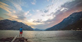 Persona su una riva rocciosa con vista sul Lago di Garda, sulle montagne circostanti e sul cielo serale dalle tonalità rossastre.