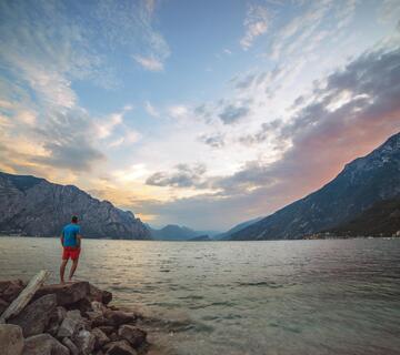 Person auf einem felsigen Ufer mit Blick auf den Gardasee, die umliegenden Berge und den rötlichen Abendhimmel.