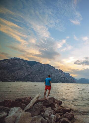 Person on a rocky shore overlooking Lake Garda, the surrounding mountains and the reddish evening sky.