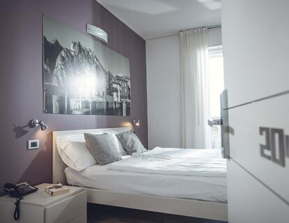 Camera d'hotel luminosa con letto matrimoniale, comodino, grande quadro in bianco e nero sopra il letto e porta della camera aperta.