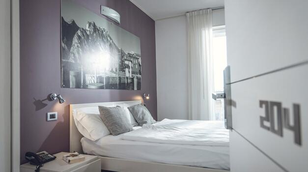 Helles Hotelzimmer mit Doppelbett, Nachttisch, großem Schwarz-Weiß-Bild über dem Bett und geöffneter Zimmertür.