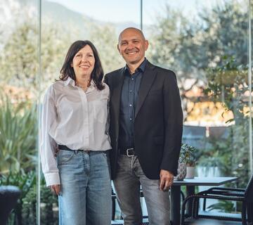 Lächelnde Frau und Mann stehen nebeneinander vor großen Glasfenstern im Hotel Villa Alberta.