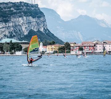 Several windsurfers with colourful sails on Lake Garda, with colourful houses and the surrounding mountain landscape in the background.