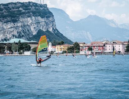 Several windsurfers with colourful sails on Lake Garda, with colourful houses and the surrounding mountain landscape in the background.