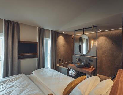 Atlas Suite with double bed, warm lighting accents and a view into the adjoining bathroom with washbasin, round mirror and shower.