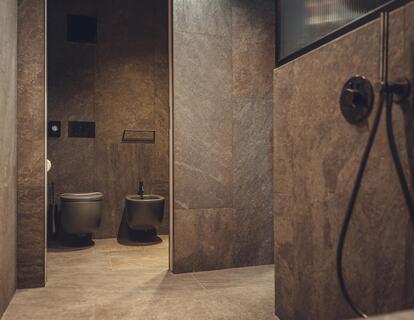 Modern bathroom with dark stone tiles, walk-in shower, black wall-mounted fixtures and a view of the toilet and bidet.