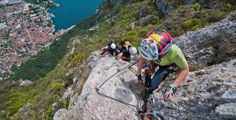 Several climbers wearing helmets and climbing gear on a steep, rocky via ferrata above Lake Garda.