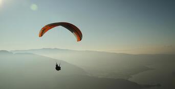 Paraglider with an orange canopy flying over a vast mountain landscape with the sun low on the horizon.