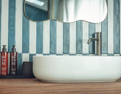 Lavabo rotondo su un piano in legno nel bagno della Suite Aura con prodotti per la cura del corpo, specchio e piastrelle bianche e blu.
