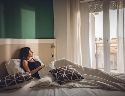 Frau sitzt auf einem Doppelbett mit weißer Bettwäsche und dunkel gemusterten Kissen in einem Hotelzimmer mit Balkon.