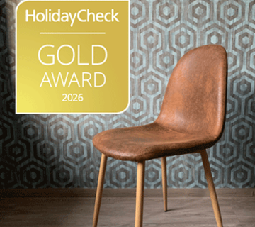 HolidayCheck Gold Award 2026 Logo vor einer gemusterten Wand mit braunem Stuhl und Holzboden.