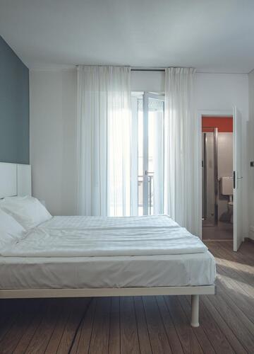 Zimmer im Hotel Villa Alberta mit Doppelbett, weißem Kopfteil, Holzboden, großem Spiegel, Schreibtisch mit schwarzem Stuhl, Blick ins Badezimmer und auf den Balkon.