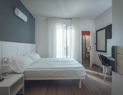 Zimmer im Hotel Villa Alberta mit Doppelbett, weißem Kopfteil, Holzboden, großem Spiegel, Schreibtisch mit schwarzem Stuhl, Blick ins Badezimmer und auf den Balkon.