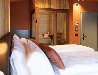 Modernes Hotelzimmer mit Doppelbett, gepolstertem Kopfteil, warmen Wandleuchten und Blick auf die angrenzende Sauna mit Glastür.