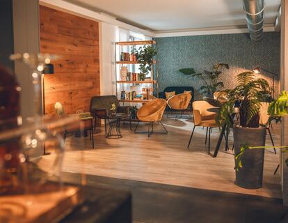 Accogliente sala comune con poltrone, tavoli, libreria e piante in un ambiente luminoso e arredato in stile moderno.